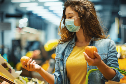 A young woman wearing a face mask and holding a shopping basket stands in a supermarket comparing two apples in each of her hands.